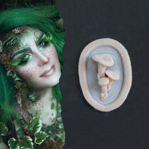 May include: A light pink mushroom mold with three mushrooms inside a light gray oval. The mold is on a dark gray background. A woman with green hair and makeup is on the left side of the image.