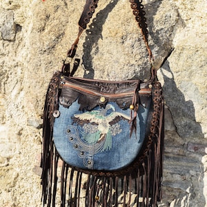 Pu&ograve; includere: Una borsa a mano in denim blu con frange in pelle marrone e una tracolla in pelle marrone con cerchi decorativi. La borsa ha un design di uccello ricamato e una chiusura a zip.