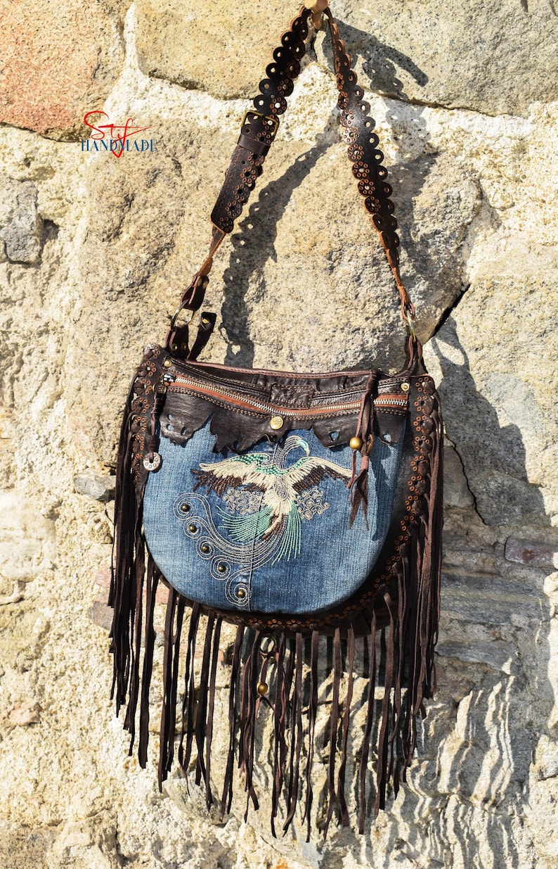 Pu&ograve; includere: Una borsa a mano in denim blu con frange in pelle marrone e una tracolla in pelle marrone con cerchi decorativi. La borsa ha un design di uccello ricamato e una chiusura a zip.