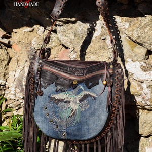 Pu&ograve; includere: Una borsa a tracolla in pelle marrone con dettagli a frange. La borsa ha una patta in denim con un design di uccello ricamato e una chiusura a zip. La tracolla &egrave; decorata con anelli metallici.