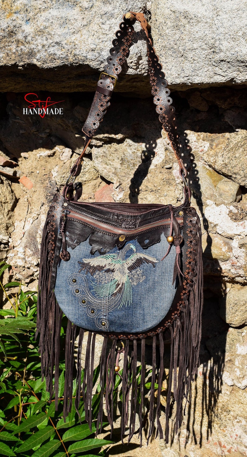 Pu&ograve; includere: Una borsa a tracolla in pelle marrone con dettagli a frange. La borsa ha una patta in denim con un design di uccello ricamato e una chiusura a zip. La tracolla &egrave; decorata con anelli metallici.