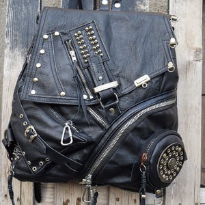 Puede incluir: Mochila de cuero negro con múltiples compartimentos con cremallera, herrajes plateados y detalles con tachuelas. Cuenta con correas ajustables y un diseño único con un detalle redondo y tachonado en el lateral. Un accesorio elegante y funcional.
