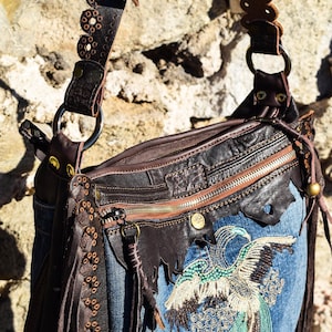 Pu&ograve; includere: Una borsa a mano in denim blu con una patta in pelle marrone e un design di uccello con perline. La borsa ha una lunga tracolla in pelle marrone con cuciture decorative e dettagli a frangia.