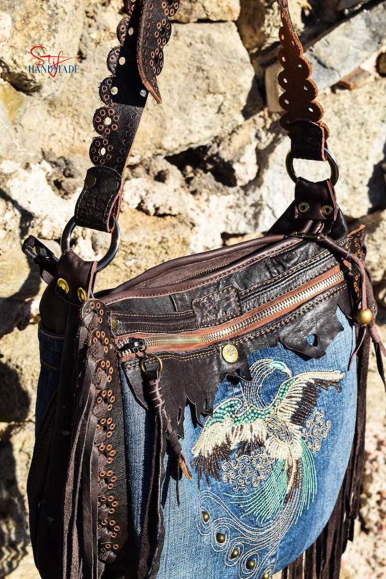 Pu&ograve; includere: Una borsa a mano in denim blu con una patta in pelle marrone e un design di uccello con perline. La borsa ha una lunga tracolla in pelle marrone con cuciture decorative e dettagli a frangia.