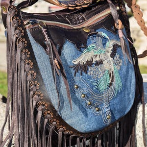 Pu&ograve; includere: Una borsa a mano in denim blu con rifiniture in pelle marrone e frange. La borsa presenta un design ricamato di un uccello con accenti turchesi e argento. La borsa ha una chiusura a zip e una tracolla in pelle marrone.