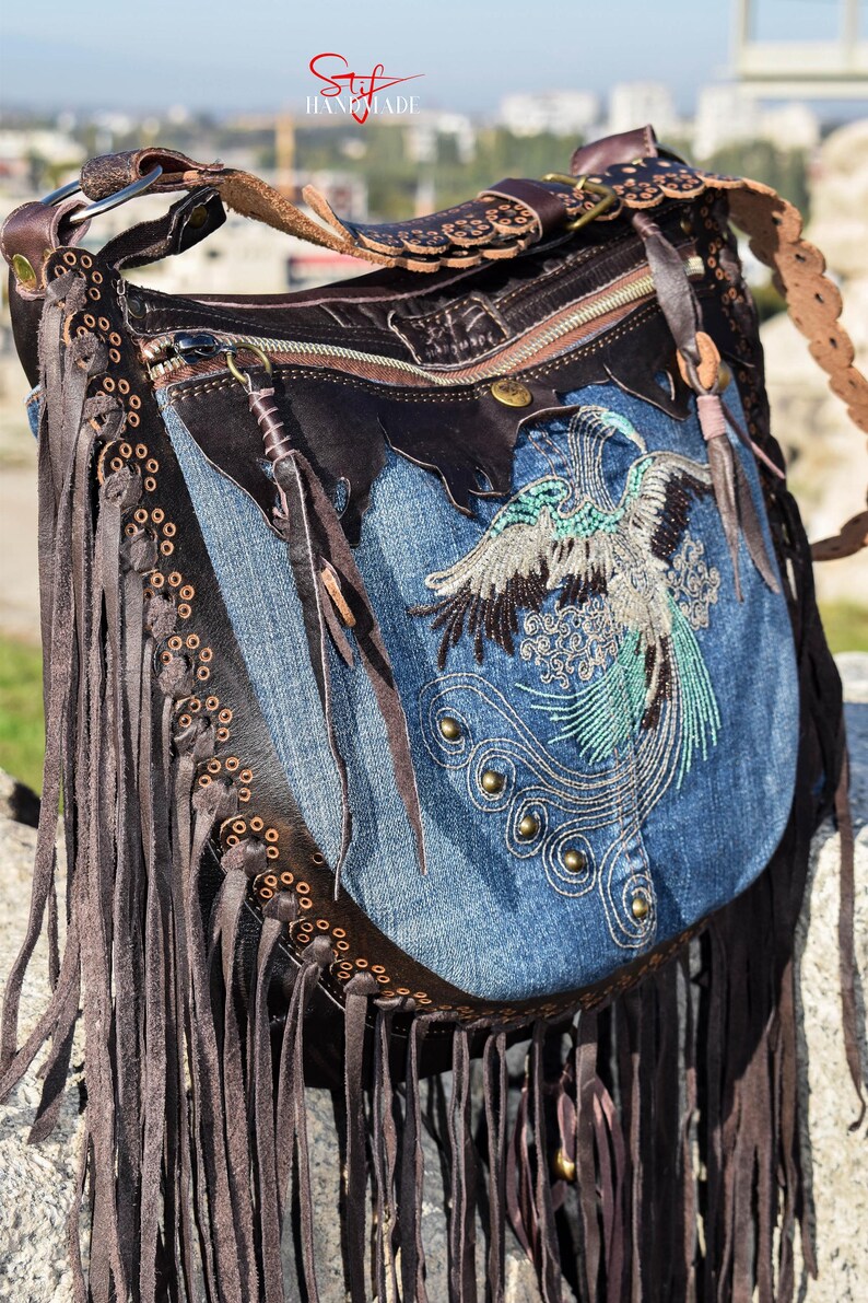 Pu&ograve; includere: Una borsa a mano in denim blu con rifiniture in pelle marrone e frange. La borsa presenta un design ricamato di un uccello con accenti turchesi e argento. La borsa ha una chiusura a zip e una tracolla in pelle marrone.