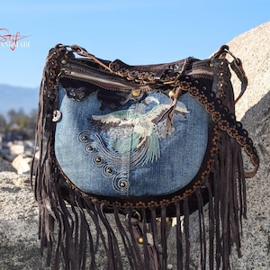 Pu&ograve; includere: Una borsa a mano in denim blu con finiture in pelle marrone e frange. La borsa presenta un design di uccello ricamato e una chiusura a zip.