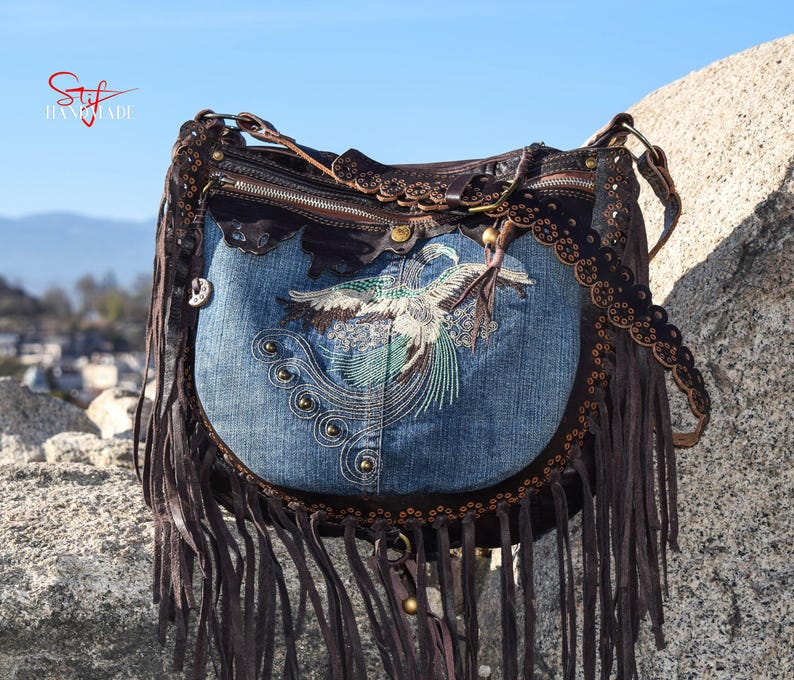 Pu&ograve; includere: Una borsa a mano in denim blu con finiture in pelle marrone e frange. La borsa presenta un design di uccello ricamato e una chiusura a zip.