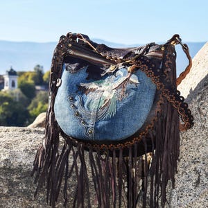 Pu&ograve; includere: Una borsa a mano in pelle marrone con una patta in denim blu con un designo di uccello ricamato. La borsa ha dettagli a frangia e una chiusura a zip.
