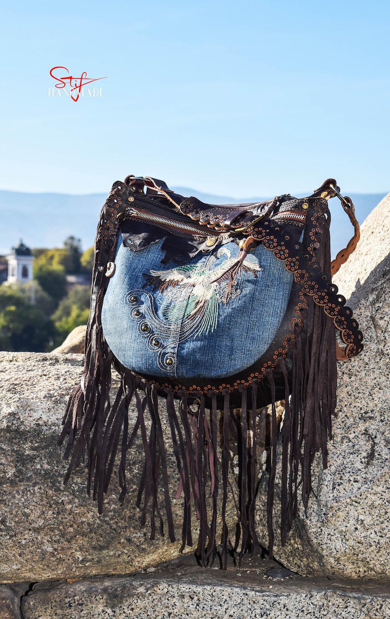 Pu&ograve; includere: Una borsa a mano in pelle marrone con una patta in denim blu con un designo di uccello ricamato. La borsa ha dettagli a frangia e una chiusura a zip.
