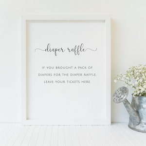 May include: A framed sign with the words "diaper raffle" in elegant script. Below, the text reads: "If you brought a pack of diapers for the diaper raffle, leave your tickets here." A metal watering can with white flowers sits to the right.