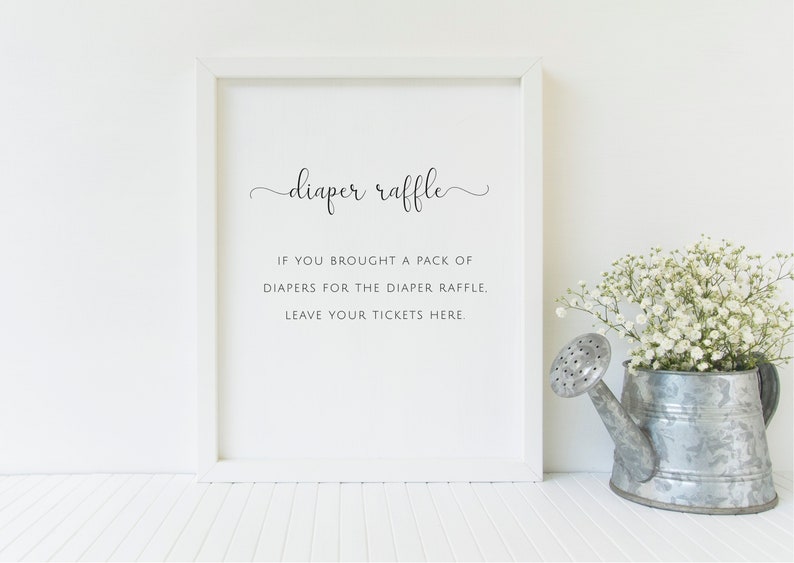 May include: A framed sign with the words "diaper raffle" in elegant script. Below, the text reads: "If you brought a pack of diapers for the diaper raffle, leave your tickets here." A metal watering can with white flowers sits to the right.