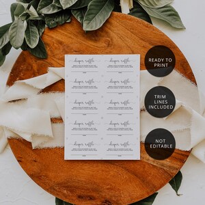 May include: A set of diaper raffle cards with the text "diaper raffle" in elegant script. The cards are on a wooden surface, surrounded by greenery and white flowers. The text "READY TO PRINT", "TRIM LINES INCLUDED", and "NOT EDITABLE" are also visible.