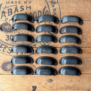 May include: A collection of fifteen dark gray metal cup drawer pulls arranged in five rows of three. Each pull features a semi-circular handle design with two small circular screw holes on the ends for mounting to a cabinet or drawer front.