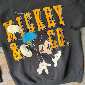 Puede incluir: Sudadera negra con un gr&aacute;fico de Mickey Mouse & Co. amarillo y blanco. Mickey Mouse est&aacute; representado en un estilo de dibujos animados azul y amarillo.