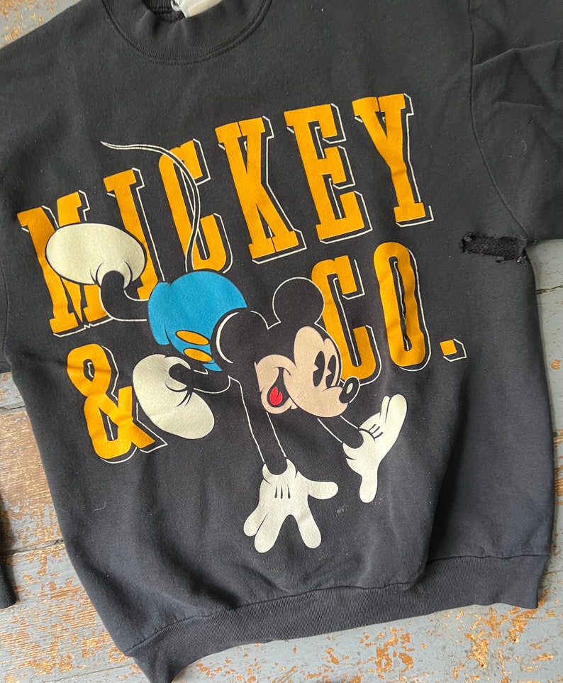 Puede incluir: Sudadera negra con un gr&aacute;fico de Mickey Mouse & Co. amarillo y blanco. Mickey Mouse est&aacute; representado en un estilo de dibujos animados azul y amarillo.