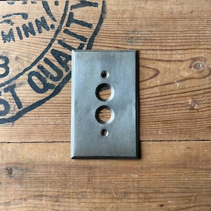 May include: A rectangular, gray metal electrical cover plate with three holes. The plate has a brushed metal finish and is set against a wooden background. The wood has a rustic appearance with visible grain and knots.