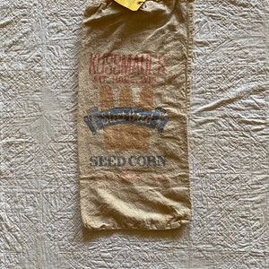 May include: A vintage burlap sack with the text "Kussmaul's Seed Corn" printed in red and blue. The sack is folded and lying on a white quilted surface.