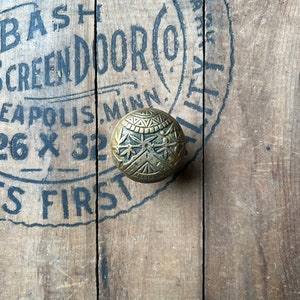 May include: A gold-toned metal door knob with a decorative design. The knob is on a wooden surface with black text that reads "Crystalized Made By Bash Screen Door (O) Minneapolis Minn. 26 x 32 S First"