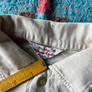 May include: Close-up of a beige polo shirt collar with a white tag that reads "70% Cotton 30% Dacron Polyester" and "M" size. The tag also includes the brand name "Cromleigh" and "Smart-Cuff" in smaller text.