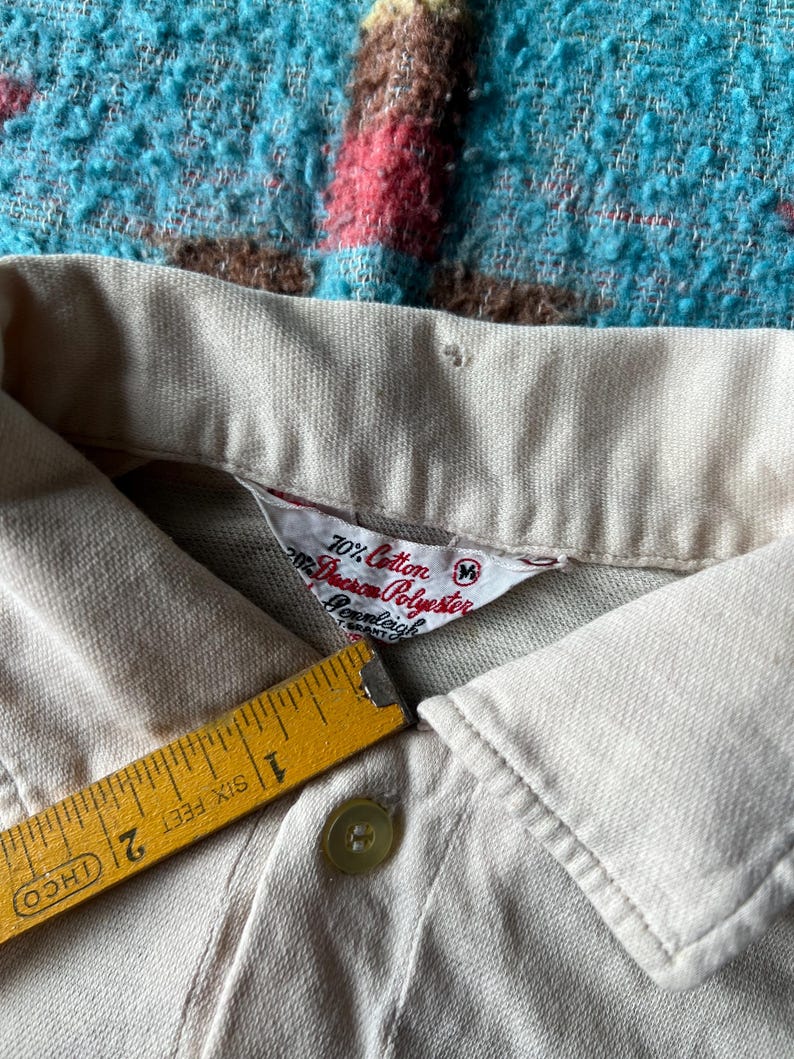 May include: Close-up of a beige polo shirt collar with a white tag that reads "70% Cotton 30% Dacron Polyester" and "M" size. The tag also includes the brand name "Cromleigh" and "Smart-Cuff" in smaller text.