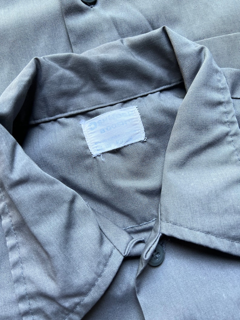 以下が含まれることがあります： 「Big Smith」と「Made in USA」と書かれた白いラベルが付いたグレーの作業着シャツです。