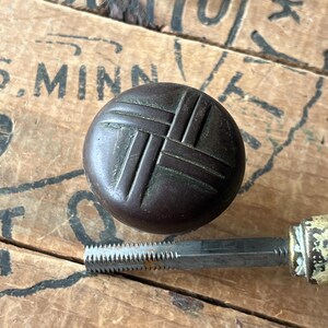 May include: A dark brown, round knob with a woven pattern, and a metal tap tool with a serrated edge. The knob and tool are set against a weathered wooden surface with faded text.