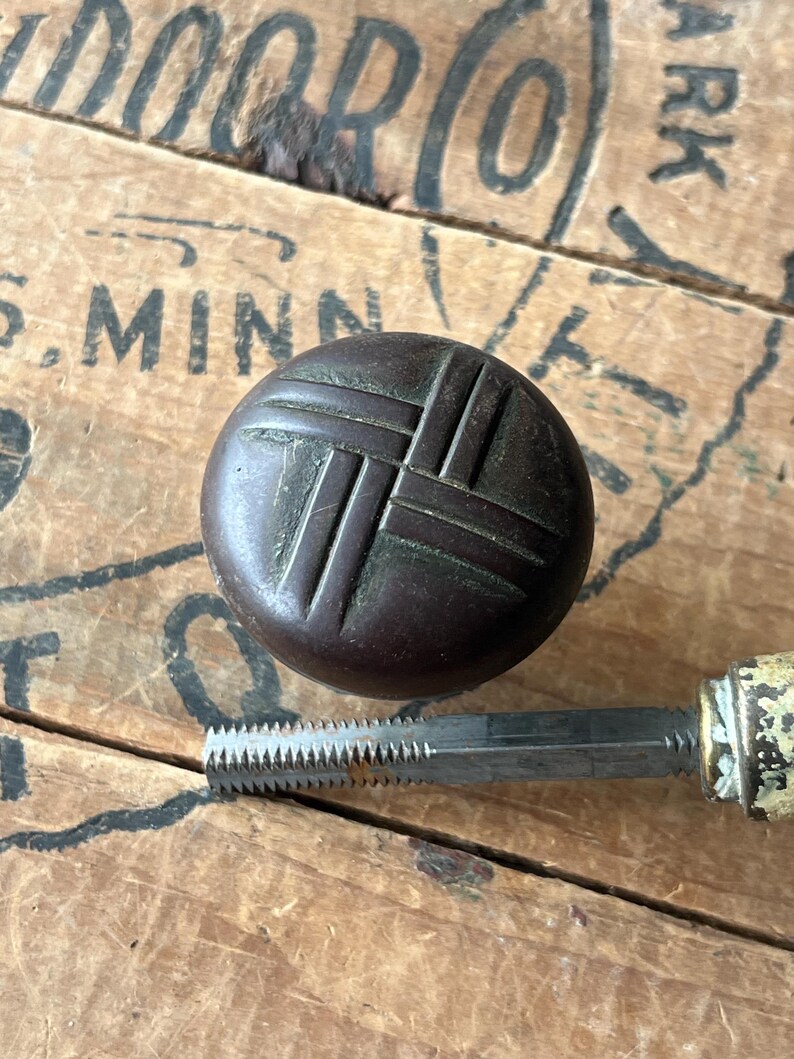 May include: A dark brown, round knob with a woven pattern, and a metal tap tool with a serrated edge. The knob and tool are set against a weathered wooden surface with faded text.