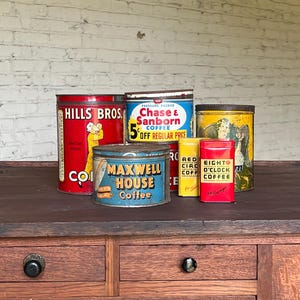 May include: A collection of vintage coffee tins, including Hills Bros, Chase & Sanborn, and Maxwell House. The tins are various sizes and colors, with labels featuring text and graphics. They are arranged on a wooden surface.