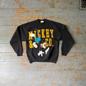 Puede incluir: Sudadera negra con un gr&aacute;fico de Mickey Mouse & Co. en amarillo y negro. Mickey Mouse est&aacute; representado en un estilo de dibujos animados, saltando y usando pantalones cortos azules.