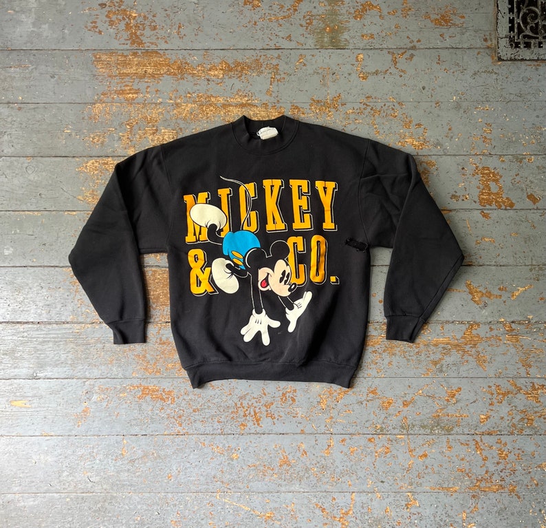 Puede incluir: Sudadera negra con un gr&aacute;fico de Mickey Mouse & Co. en amarillo y negro. Mickey Mouse est&aacute; representado en un estilo de dibujos animados, saltando y usando pantalones cortos azules.