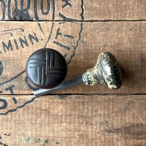 May include: Two antique door knobs. One is a dark brown, round knob with a crosshatch design. The other is an off-white and black knob with a decorative pattern. Both knobs are attached to a metal spindle.