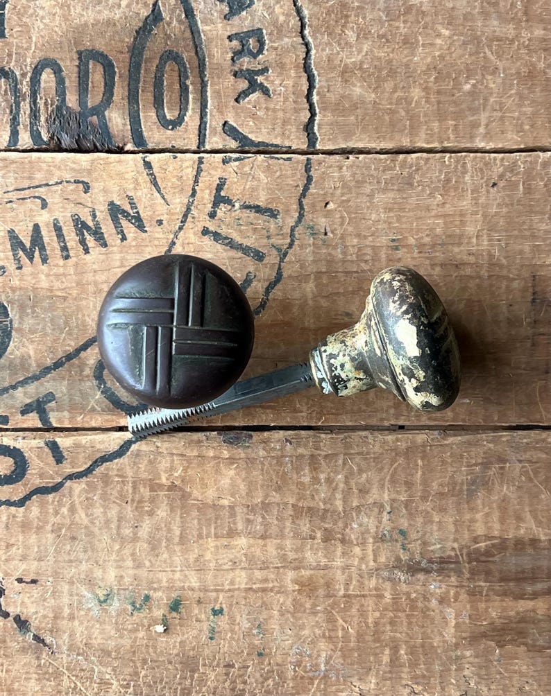 May include: Two antique door knobs. One is a dark brown, round knob with a crosshatch design. The other is an off-white and black knob with a decorative pattern. Both knobs are attached to a metal spindle.
