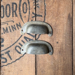 May include: Two antique silver drawer pulls with a rounded, slightly curved shape. The pulls are set against a wooden background.