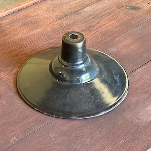 May include: A black metal lamp shade with a circular base and a cylindrical top. The shade has a glossy finish and shows signs of wear. The object is set against a wooden surface.