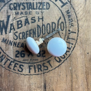 May include: Two white, oval-shaped door knobs with metal hardware, resting on a weathered wooden surface. The knobs are positioned near a partially visible, circular stamp with the text "WABASH SCREEN DOOR". The wood has a rustic, aged appearance.