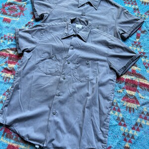 以下が含まれることがあります： ポケット付きのグレーの半袖ボタンダウンシャツが2枚、重ねて置かれています。シャツは青と茶色の模様の毛布の上に置かれています。