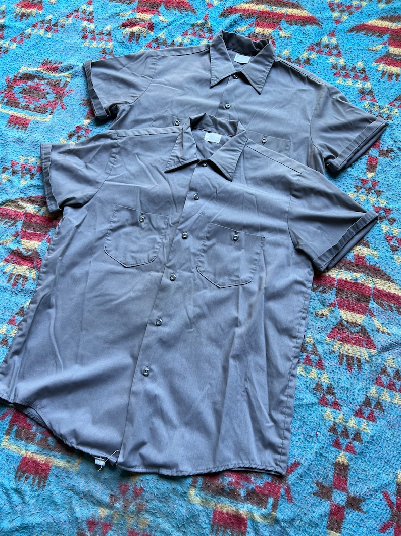 以下が含まれることがあります： ポケット付きのグレーの半袖ボタンダウンシャツが2枚、重ねて置かれています。シャツは青と茶色の模様の毛布の上に置かれています。