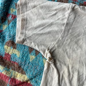 May include: Close-up of the beige fabric of a t-shirt sleeve. The fabric is slightly wrinkled and has a few small holes.
