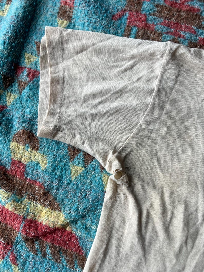 May include: Close-up of the beige fabric of a t-shirt sleeve. The fabric is slightly wrinkled and has a few small holes.