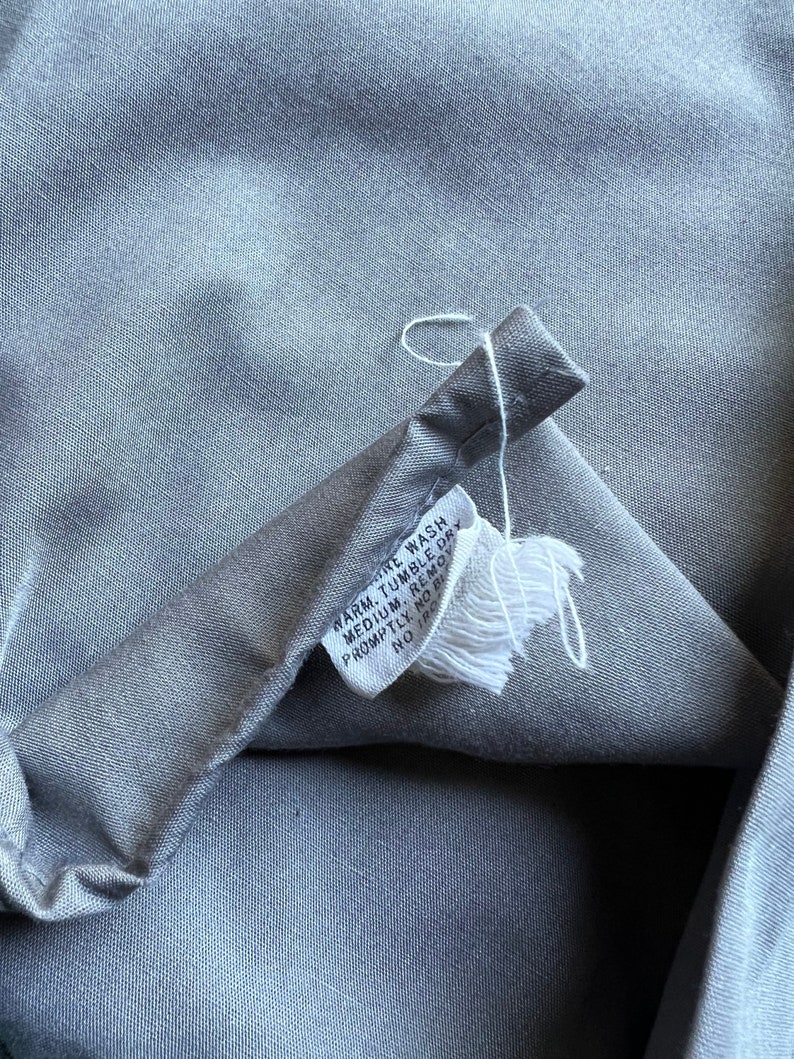 以下が含まれることがあります： グレーの布地のお手入れラベルに、白い文字で「温水で洗濯機洗い。中温でタンブラー乾燥。すぐに取り出す。漂白剤不可。アイロン不可。」と書いてあります