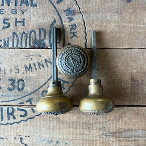 Puede incluir: Pomos de puerta antiguos de latón y una pieza central con el texto "BOARD OF EDUCATION CITY OF CHICAGO". Los pomos tienen forma redonda con base decorativa y varilla roscada. Los artículos están sobre una superficie de madera.