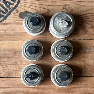 May include: Six vintage metal jar lids with black knobs. The lids are silver with white rims and embossed text. The lids are arranged on a wooden surface with a printed logo.