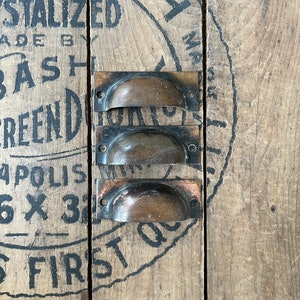 May include: Three antique brass cabinet pulls with a rounded, cup-shaped design. The pulls are arranged vertically on a wooden surface with black painted text.