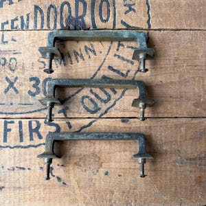 May include: Three rectangular metal drawer pulls with a weathered, dark green patina. Each handle features a horizontal bar, two square mounting plates, and two downward-facing threaded bolts with small circular nuts attached to the ends.