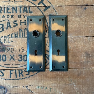 May include: Two antique door plates with a black finish and a copper patina. The plates have a rectangular shape with rounded corners and a keyhole in the center. The plates are set against a wooden background.