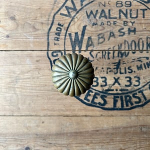 May include: A brass knob with a fluted design. The knob is centered on a wooden surface with a black and white stamp that reads "N89 WALNUT MADE BY WABASH SCREENDOOR INDIANAPOLIS, MIN 33 X 33 EES FIRST".