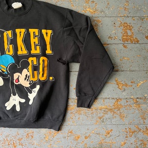 Puede incluir: Sudadera negra con un gr&aacute;fico de Mickey Mouse y el texto "Mickey Co." en letras amarillas.