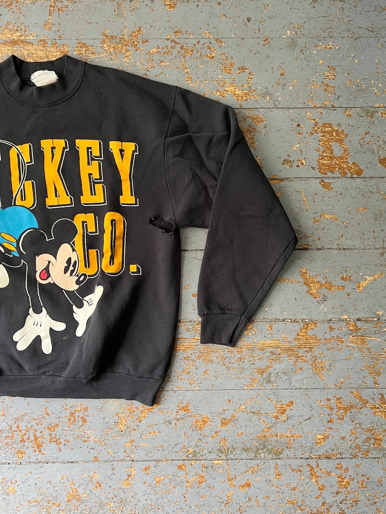 Puede incluir: Sudadera negra con un gr&aacute;fico de Mickey Mouse y el texto "Mickey Co." en letras amarillas.