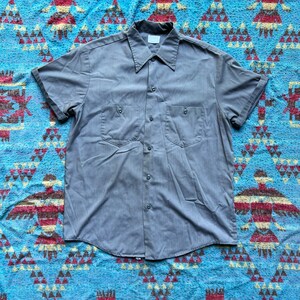 以下が含まれることがあります： ボタンダウンカラーと胸ポケットが2つ付いたグレーの半袖ワークシャツです。